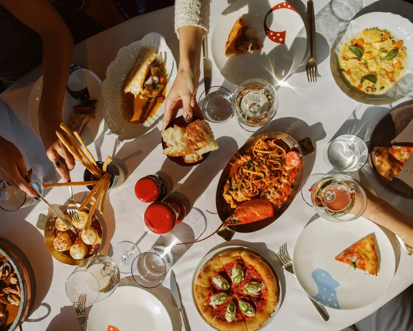 A beautifully set dining table filled with an array of gourmet dishes, captured from a top-down perspective. Guests are engaged in their meal, cutting into seared scallops, breaking fresh bread, and toasting with glasses of wine. The spread includes a rich lobster pasta, a personal-sized pizza topped with burrata and jalapeños, a seafood dish in broth, creamy ravioli with vegetables, and a serving of mussels and clams. Crispy breadsticks stand in a glass, accompanied by red and black pepper mills. Sunlight casts warm highlights on the dishes, creating an inviting and elegant dining ambiance