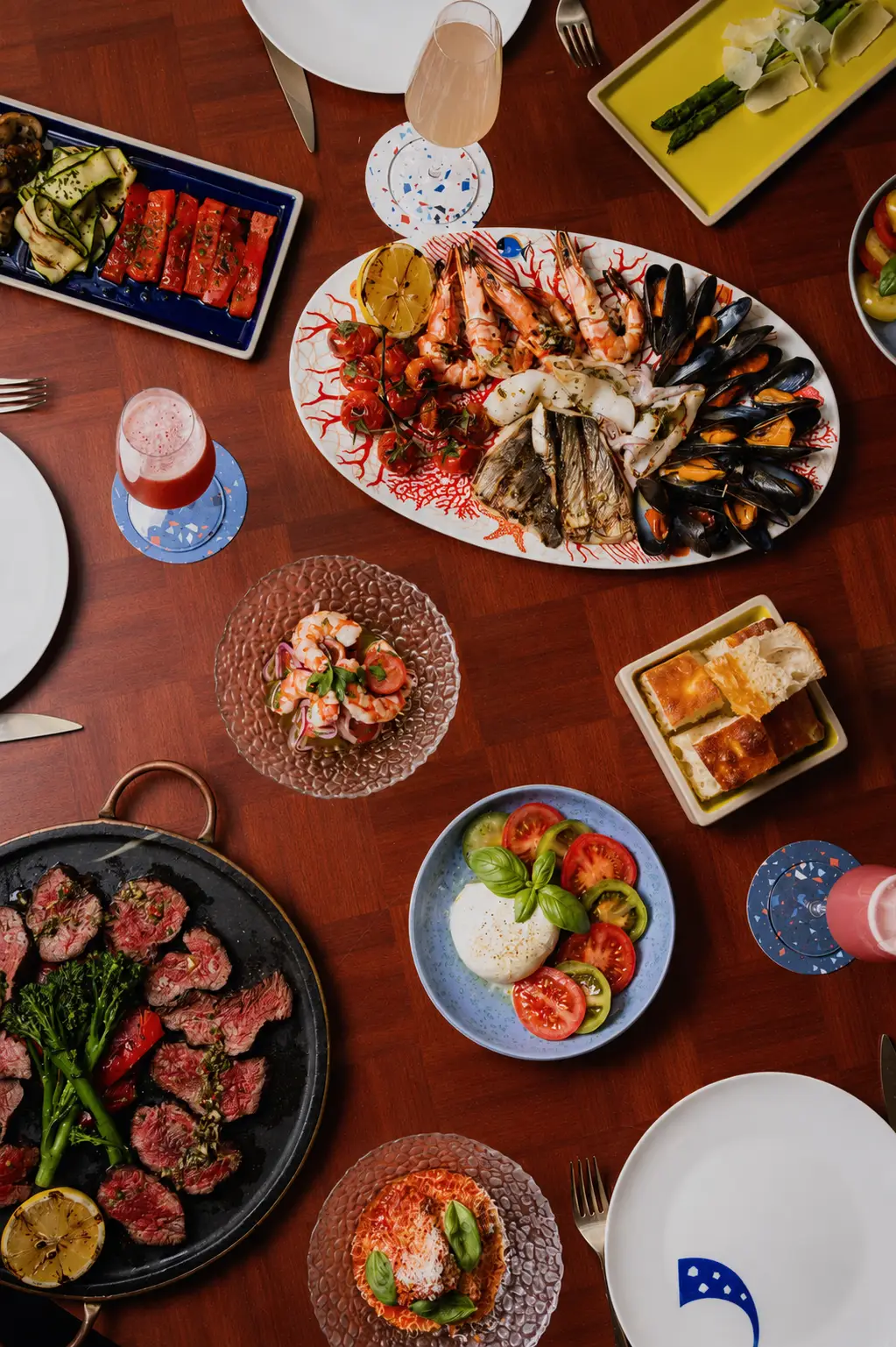 Father's Day Il Pranzo lunch at Il Gattopardo Mayfair. Food spread on a wooden table, featuring a grill plate with thinly sliced marinated beef, a platter of seafood including prawns and mussels, plates of fresh vegetables and tomatoes, and lots of side dishes and drinks.